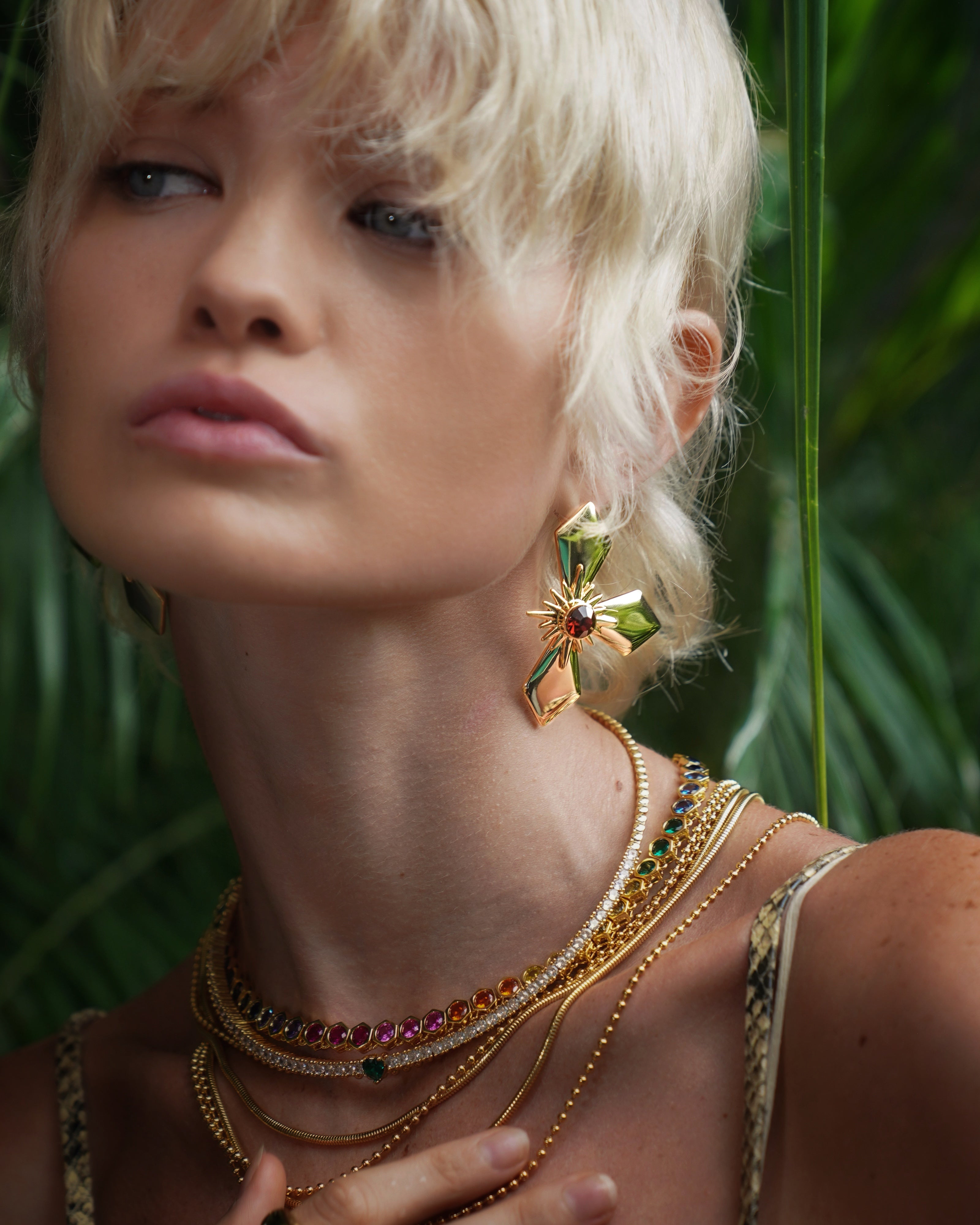 Woman with blonde hair wearing gold jewelry and large earrings in a forest setting.