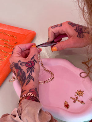 Person with tattoos working on jewelry, including cutting a chain and arranging small gold charms.