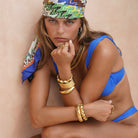 Woman wearing a blue bikini and colorful headscarf with gold jewelry against a beige background