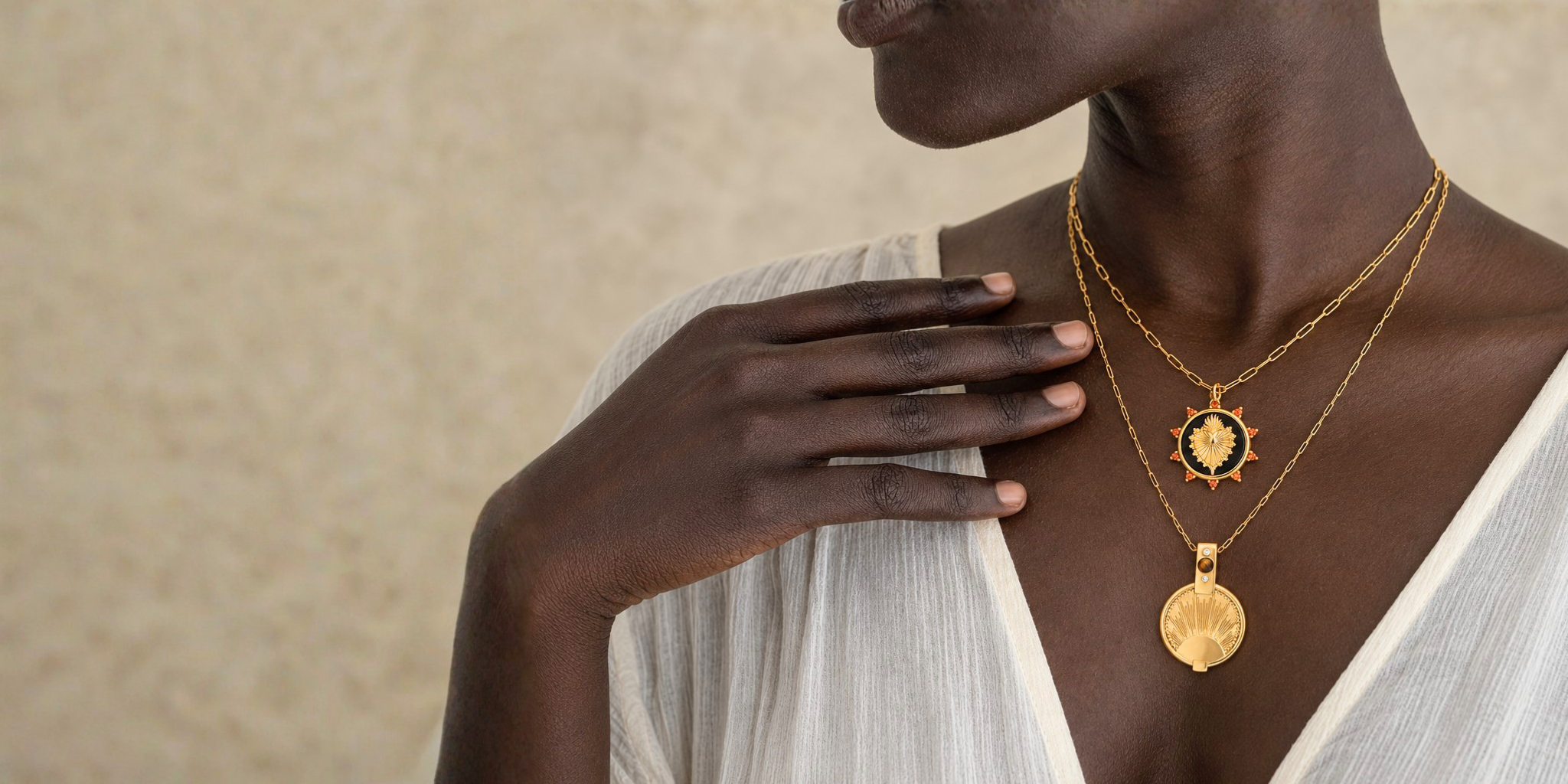 A close-up lifestyle shot of a model wearing two gold necklaces: a sunburst pendant necklace and a scallop shell pendant necklace, paired with a white draped top against a textured beige background.